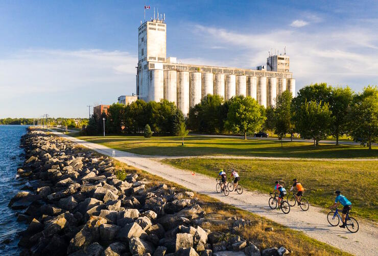 Group of cyclists riding on a road by water towards a large building.