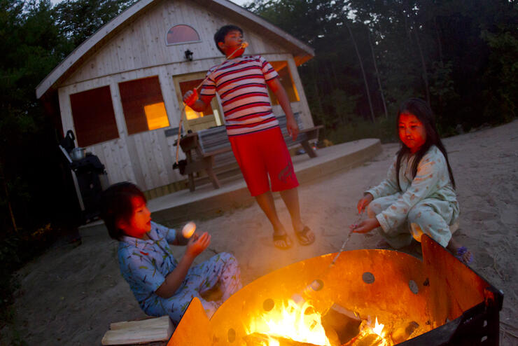 Three children roasting marshmellows over a campfire in front of a small cabin