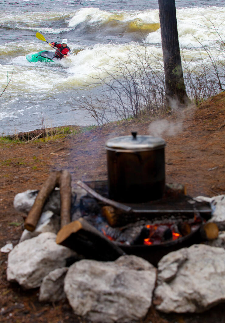 Steaming pot over campfire with a kayaker playing in whitewater in background