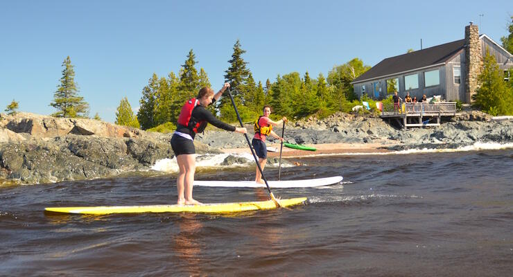 Two women paddleboarding in front of a small lodge