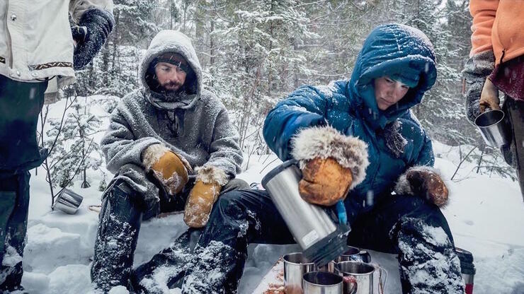 Man and woman in heavy winter clothing sitting at snowy campsite. 
