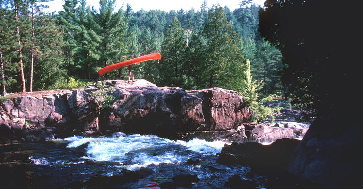 Bill Mason portaging a red canoe around rapids in Algonquin Park