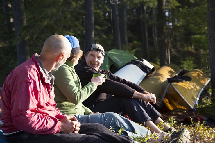 Three people sitting on ground with canoes and tent in background