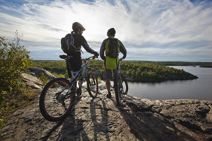 Two offroad cyclists stand with their bicycles on a rocky ridge overlooking a lake