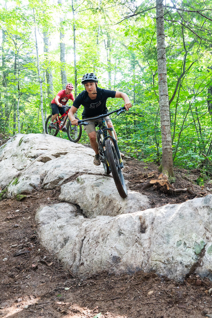 Two teenage boys ride mountain bikes over a rocky ridge in the forest