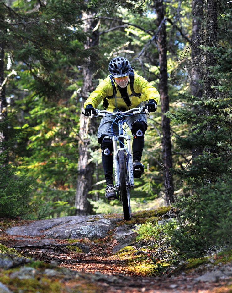 Man in yellow hi-vis jacket mountain bikes over mossy rocks in a forest