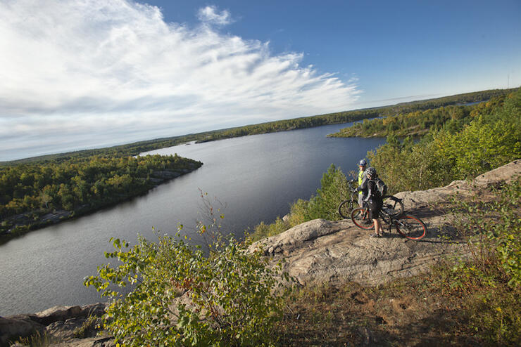 Two cyclists stop on a rocky outlook to enjoy the view of forest and lake