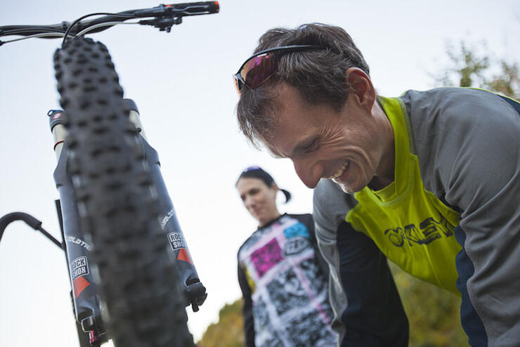 A man leans down and works on fixing a mountain bike while another cyclist looks on