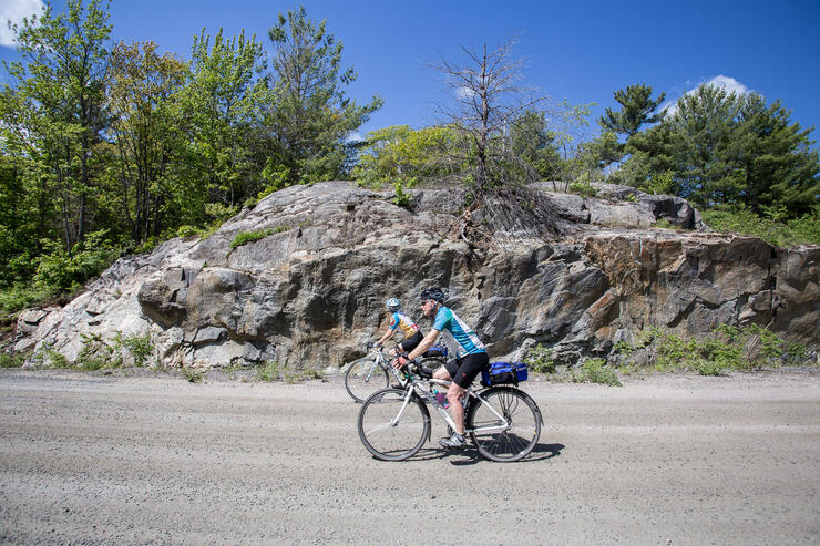 two touring cyclists ride down a road past a rock cut