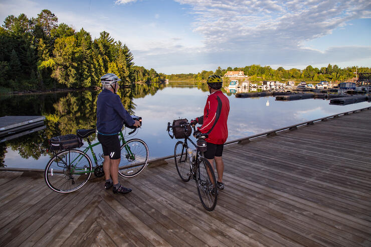 Two cyclists stand with their bikes on a boardwalk overlooking the water