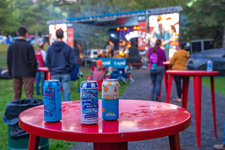 three cans of local craft beer sit on a red table while people watch a performance on stage behind