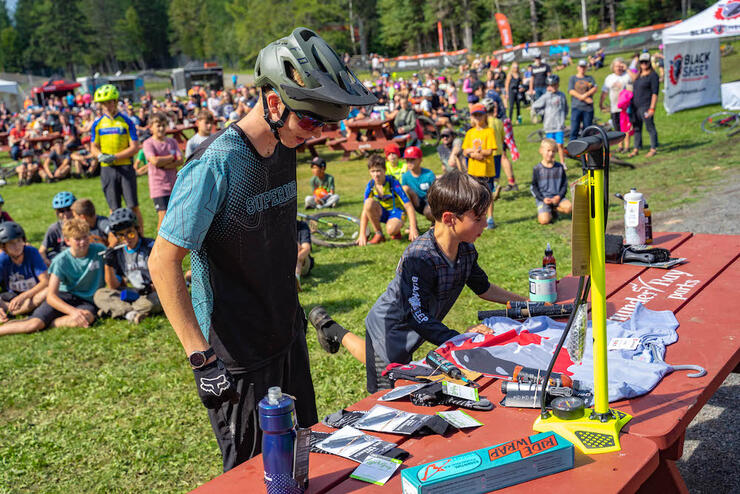 two younger cyclists look at a table with race prizes while a crowd sits on the grass behind them