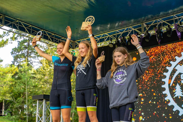 three girls wave on a stage with their prizes after a bike race