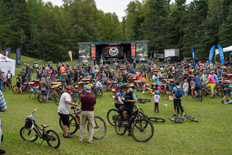 a crowd of cyclists gather in front of a stage in the grass surrounded by trees
