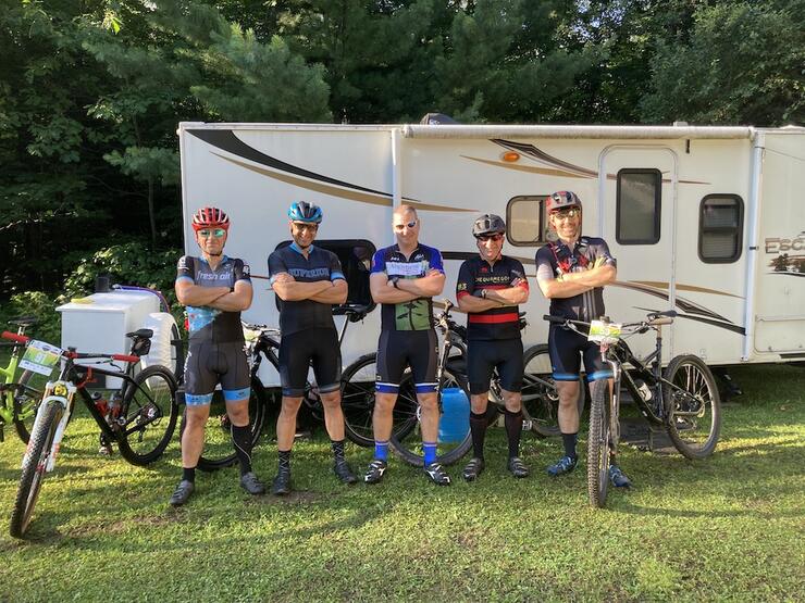 five male cyclists in racing gear pose with arms crossed in front of a trailer