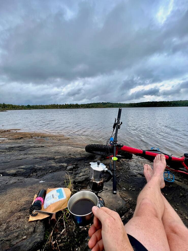 person sits on the shore with bike and camp coffee at Lake Laurentian Conservation Area in Sudbury