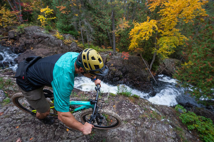 man rides a bike along the edge of a rocky outcropping looking down on flowing water in the fall in the Algoma Highlands