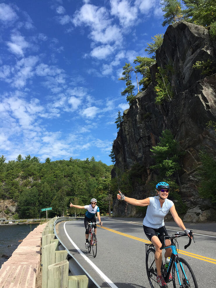 two women give thumbs up while riding past a cliff on a bicycle tour