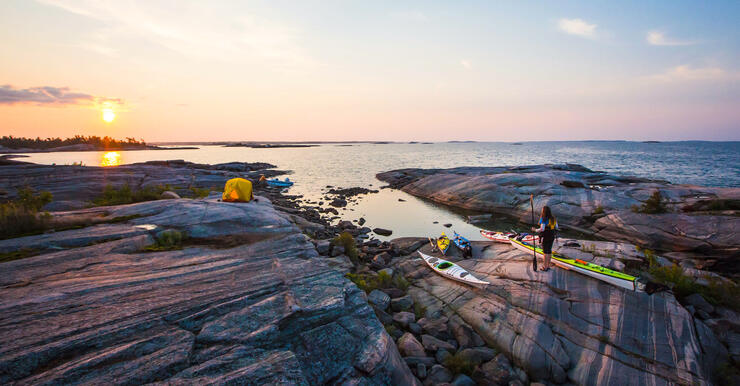 Woman standing beside kayaks on smooth rocks watching sunset 