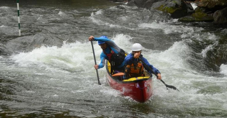 Two people with helmets and lifejackets in a red canoe paddling in whitewater rapids on Gull River. 