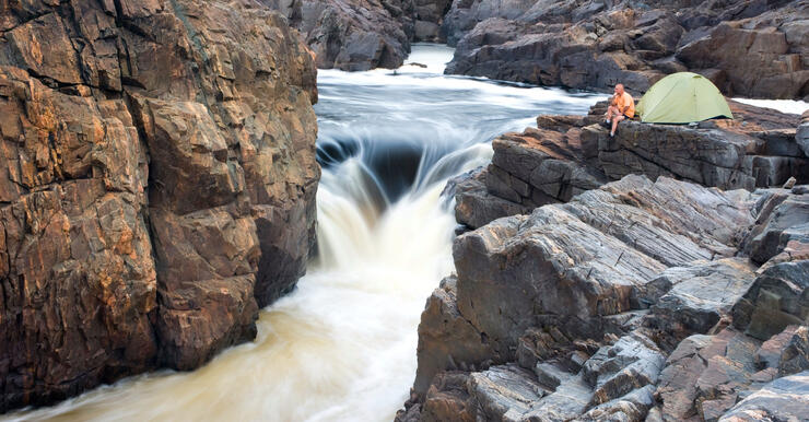 Man sitting beside green tent perched high above thundering waterfalls. 