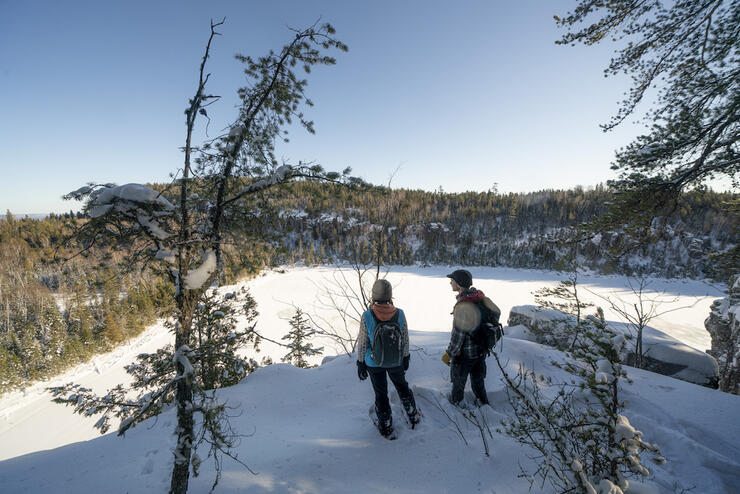 Two people on snowshoes stand atop a snowy lookout over a frozen lake