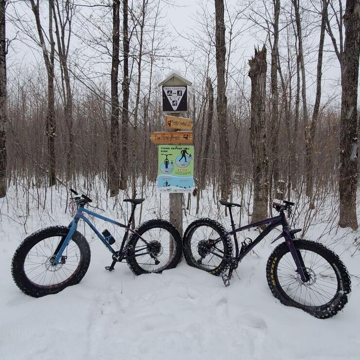 Fat bikes sitting in the snow next to a trail sign in the woods