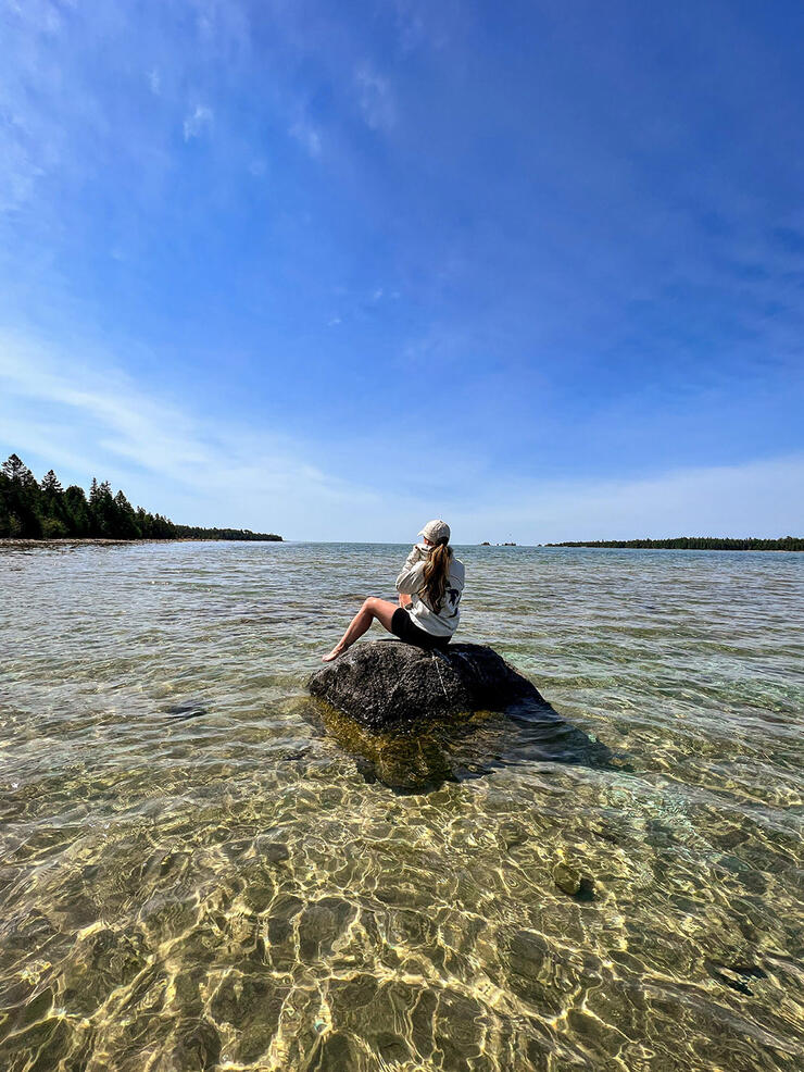 Woman sits on rock in shallow, clear water at Misery Bay Provincial Park