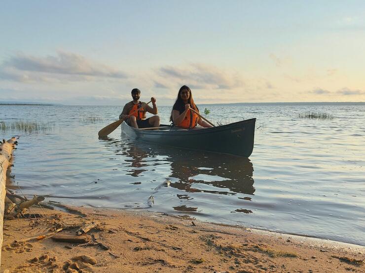 Two people paddle canoe next to sandy shore.