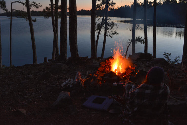Silhouette of person sitting beside campfire next to lake.