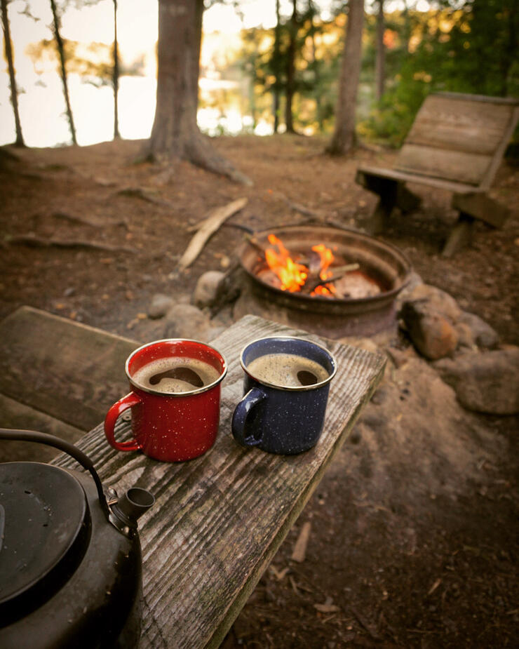 Two mugs of hot chocolate with kettle on bench next to fire on a campsite.