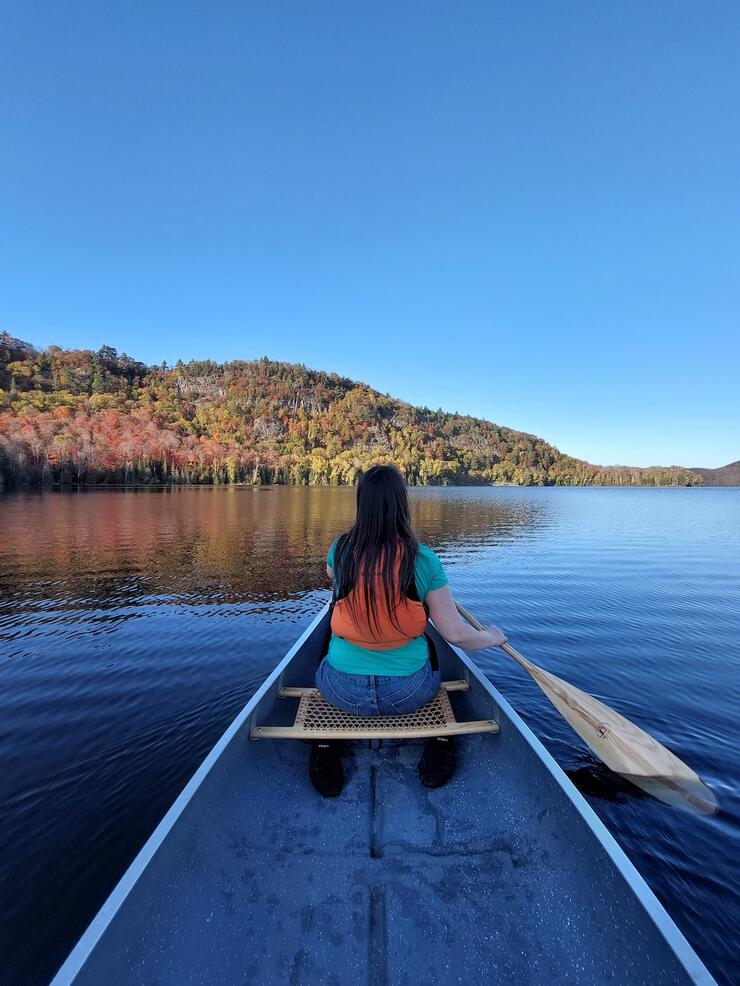View from stern of canoe of person paddling with most of leaves off the trees on shore.