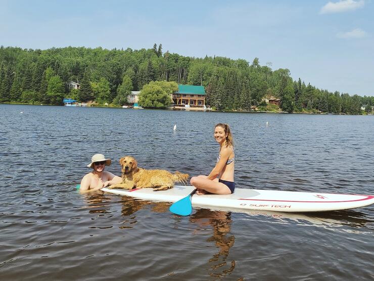 Woman sits on paddleboard with dog on a lake while man stands in water nearby