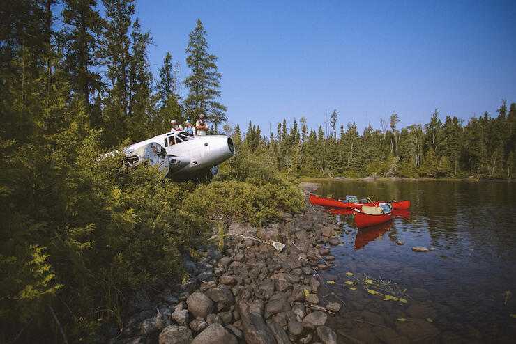 People standing on a crashed plane on shore with canoes pulled up beside.
