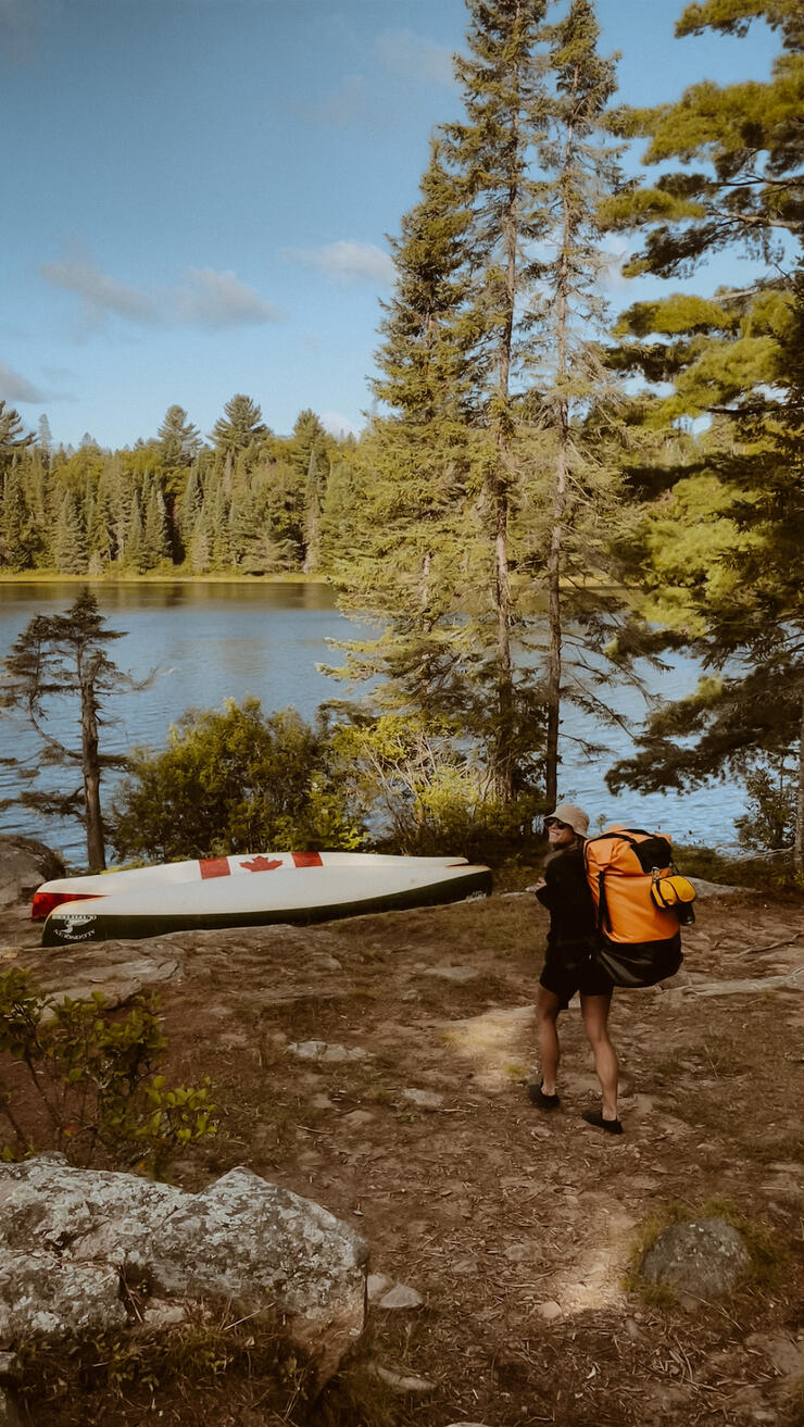 Woman walks with pack toward overturned canoes on a campsite.