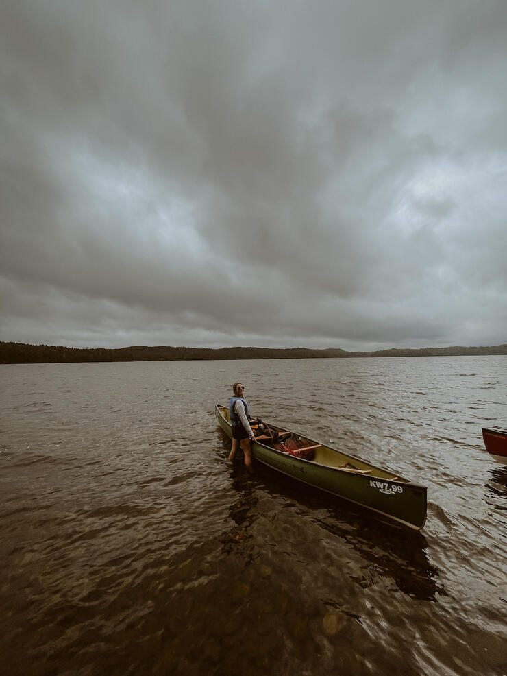 Dark clouds over lake as woman stands in water next to canoe.