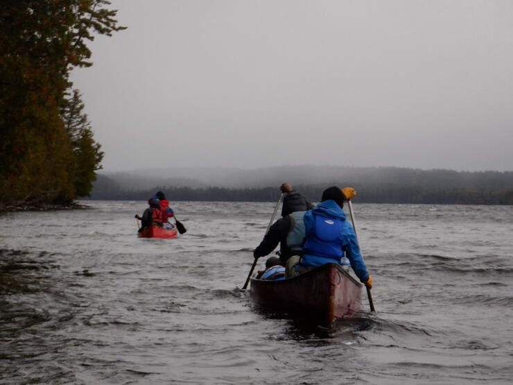 Two canoes paddle on wavy lake next to shoreline on rainy day.