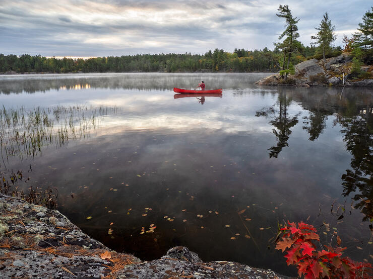 Soloist in red canoe paddles out from rocky shore across placid lake.