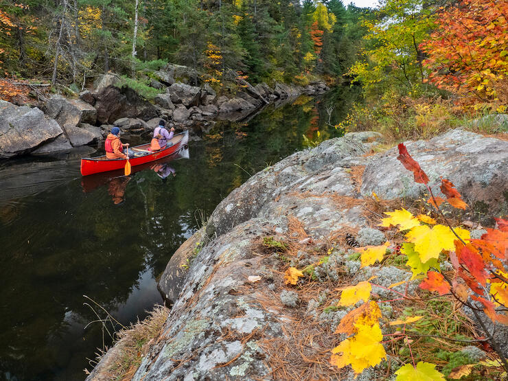 Couple in a red canoe paddles narrow passage between rocks with fall colours on the surrounding trees.