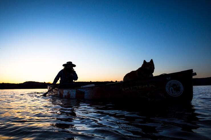 Silhouette of a man paddling a canoe with a dog in front of canoe.