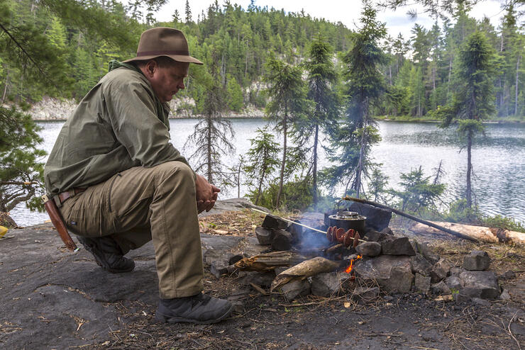 Man backcountry camping on crown land in Ontario cooking meat over a campfire at a campsite.