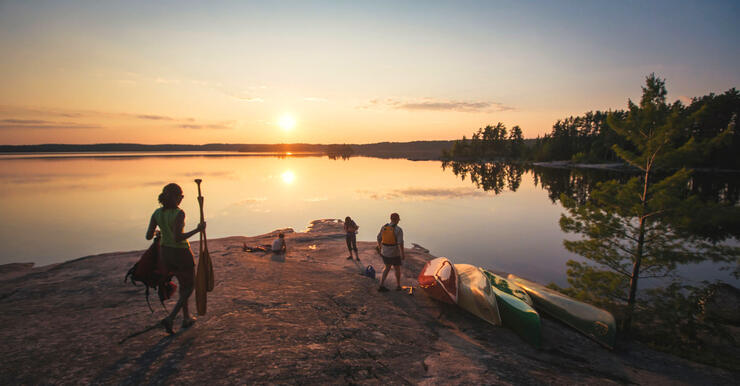 Young woman with paddles walking on rocky point towards friends watching sunset. 