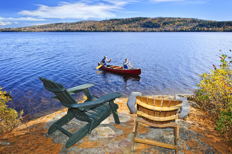 Muskoka chairs on a rock with people in canoe paddling by.