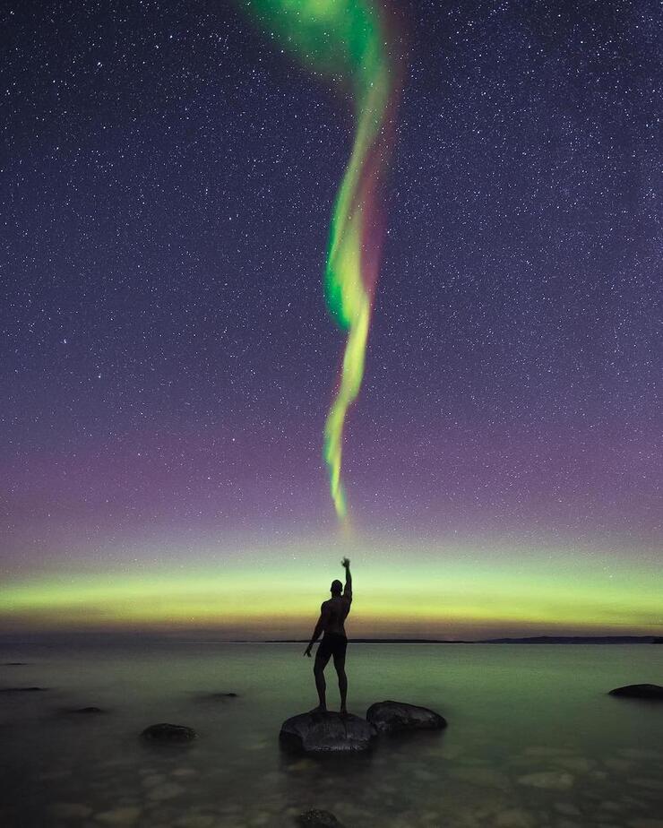 Person standing on a rock near lake shore and reaching up to touch Northern Lights in sky