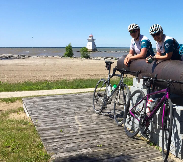 Two cyclists standing beside bicycles with beach and lighthouse in background.