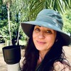 a smiling Jenn Smith Nelson, with long dark hair and a wide brimmed hat, holding up a large camera in front of palm trees and a beach.
